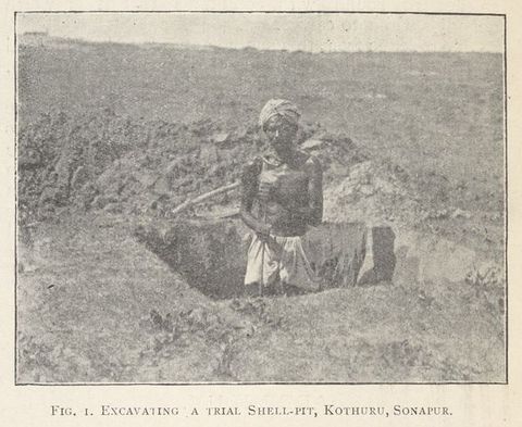 Black and white photograph. A man stands hip deep in a square hole dug into the ground. He has dark skin and wears a white turban and white shorts.