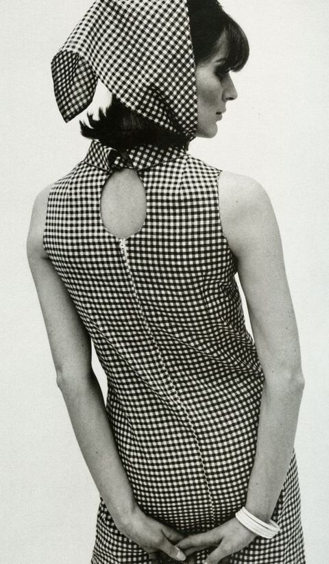 Black and white photograph. Shown from the thighs up, a woman facing away from us wears a checkered sleeveless dress with a keyhole below the high collar. A matching scarf over her dark hair is tied under her chin.