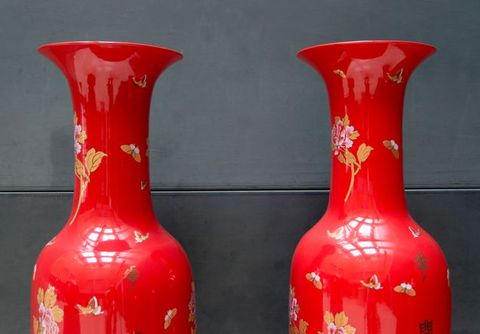 Installation photograph. The vibrant red shoulders, long necks, and flaring mouths of two ceramic vessels reflect the light in the room and the photographer taking this picture. Butterflies and flowers in shades of golden tan, white, and pale pink are scattered across the vessels’ surfaces.